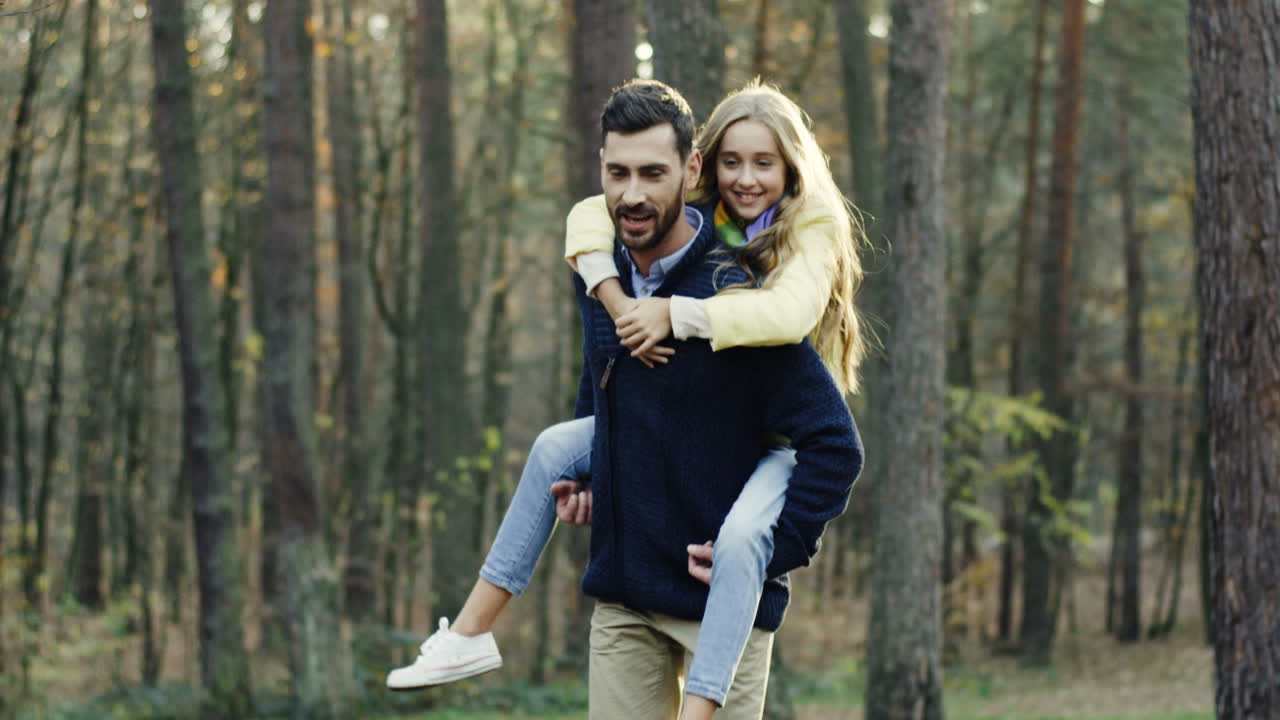 Caucasian father walking in the forest and carrying his cute small daughter on his back while they talking