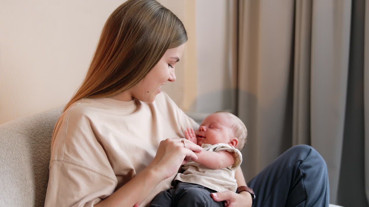 Woman with long blonde hair holding her baby. Mother takes tiny hand of a child and smiles to a kid.