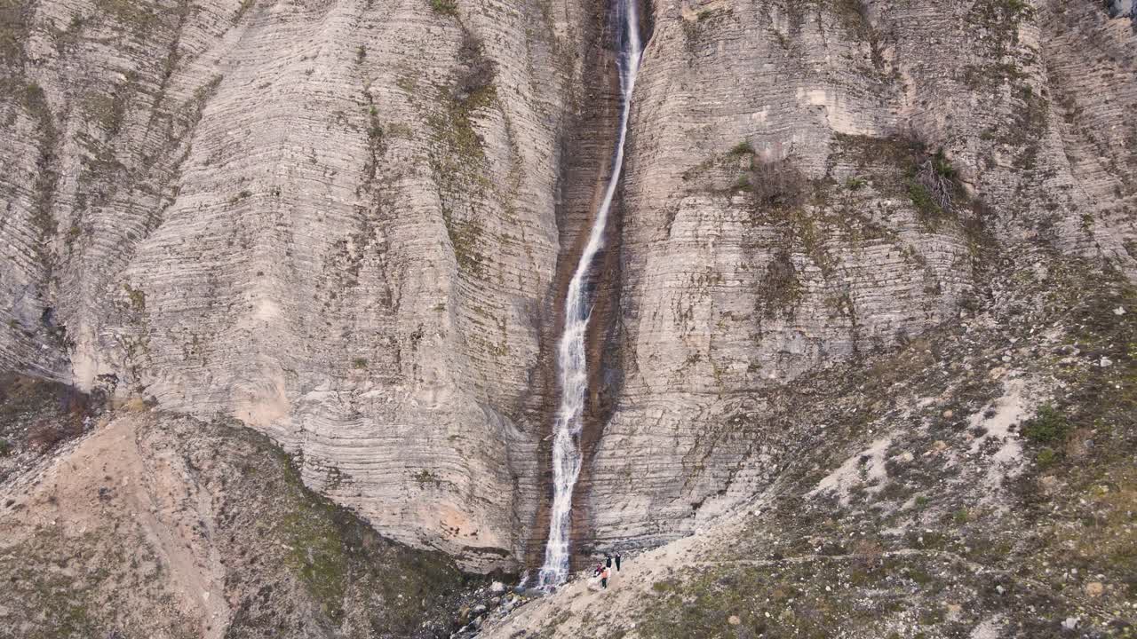 Ripresa aerea con tilt verso il basso e risalita sopra una cascata alta e sottile con persone in piedi sotto a Tzoumerka, Grecia.