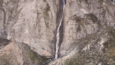 Aerial-tilt-down-and-rising-over-a-high-thin-waterfall-with-people-standing-below-in-Tzoumerka,-Greece