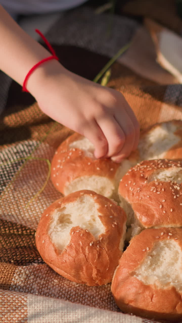 Close-up view of two young boys, faces not visible,pinching and eating bread from a cluster of rolls on a checkered picnic blanket. The scene captures a quiet, intimate moment of sharing food outdoors