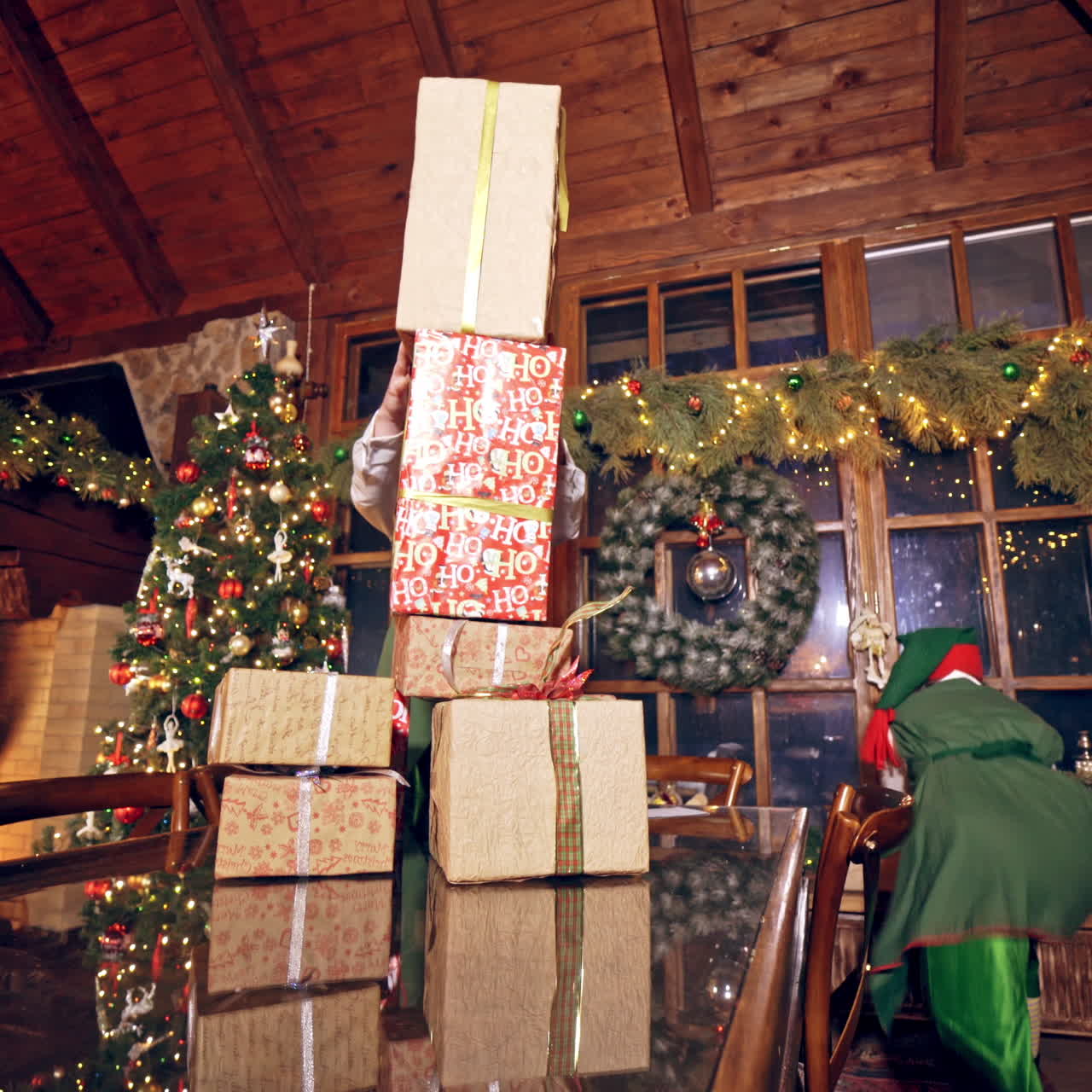 Two elves are standing in specially decorated room near the table, making a piramide from the presents. Feast fireplace in the background. Christmas spirit concept close up video. Christmas eve. New Year is coming. Elves are putting presents on the table. Presents are falling down from the table. El