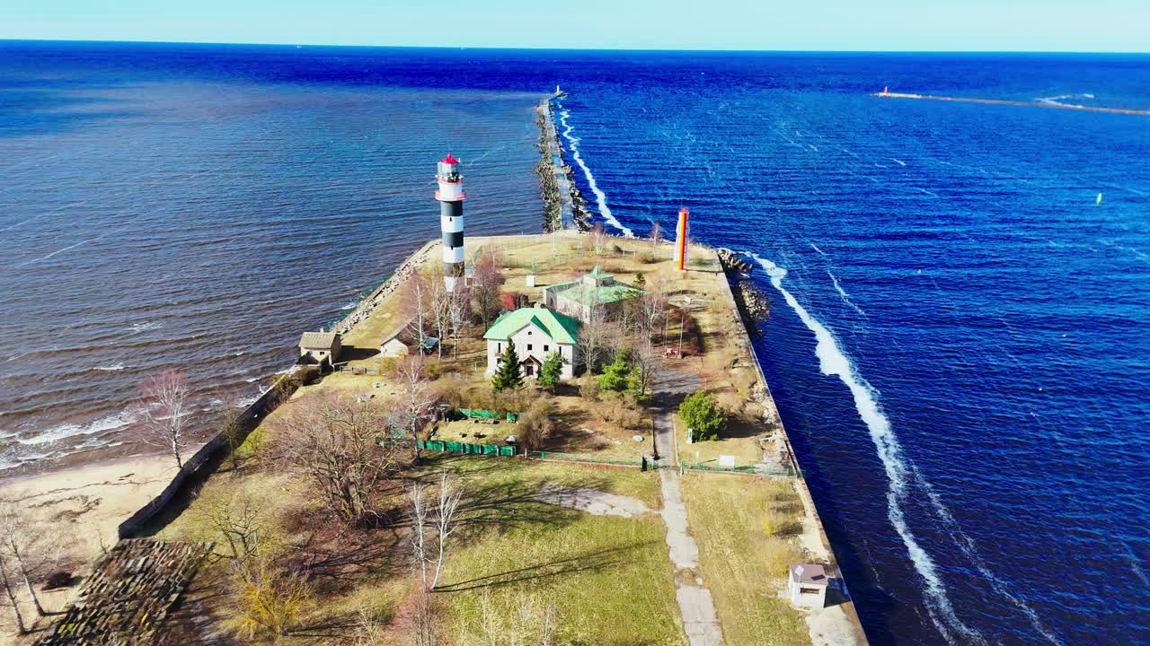 A black-and-white striped lighthouse stands at the end of a narrow jetty flanked by waves, small buildings, and sparse trees, marking the division of two bodies of water.
