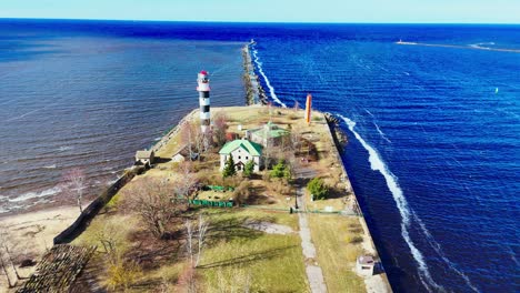 a black-and-white striped lighthouse stands at the end of a narrow jetty flanked by waves, small buildings, and sparse trees, marking the division of two bodies of water.