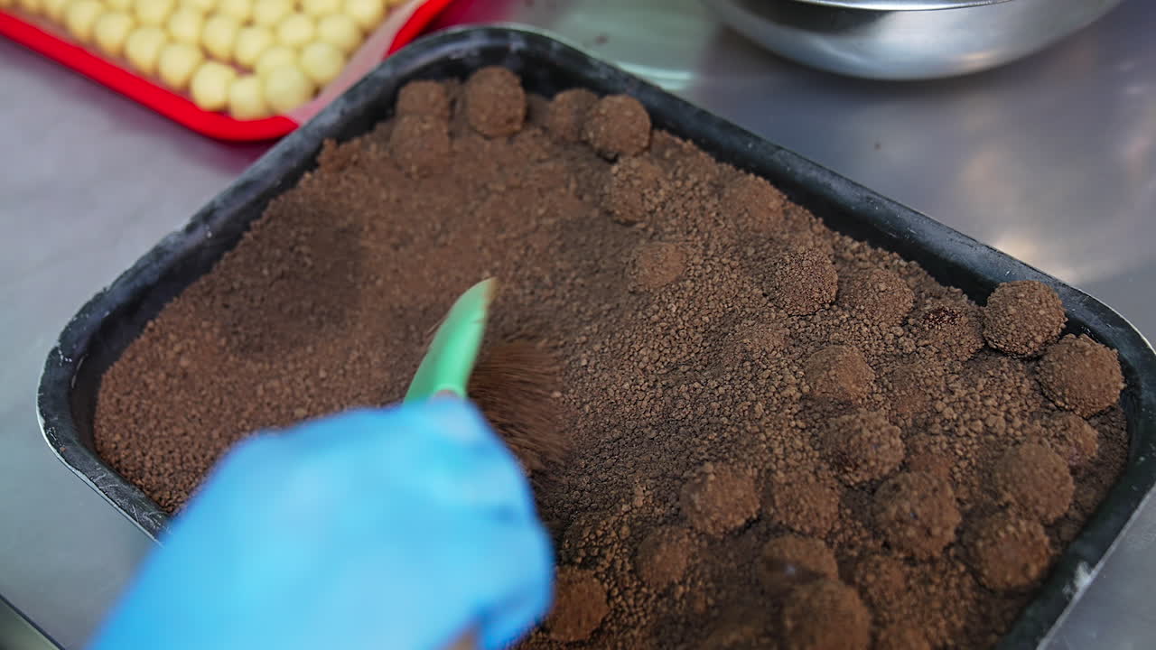 Sweet balls covered with melted chocolate thrown into tray with cocoa sprinkling. Hand holding spatula mixes candies with coating. Close up.