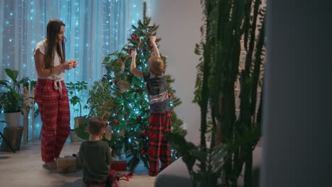 Una-Madre-De-Familia-Y-Dos-Hijos-Decoran-Un-árbol-De-Navidad-Con-Juguetes-En-El-Salón-De-Su-Casa.-Adornos-Navideños-Y-Preparación-Para-Las-Vacaciones.-Imágenes-4k-De-Alta-Calidad