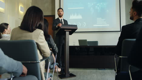 zoom de cámara sobre un hombre de negocios caucásico hablando en un podio en una sala de conferencias y mostrando algunos gráficos y gráficos en la pantalla grande