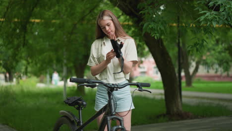 joven ajusta guantes negros mientras está de pie junto a su bicicleta en un camino pavimentado, rodeada de árboles verdes exuberantes, lleva un reloj inteligente en la mano derecha, con una vista borrosa de los edificios en el fondo