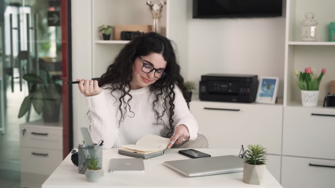 The young brunette woman with curly long hair is sitting at a desk in the office and writing something in her journal. The woman is contemplating how to write it better.