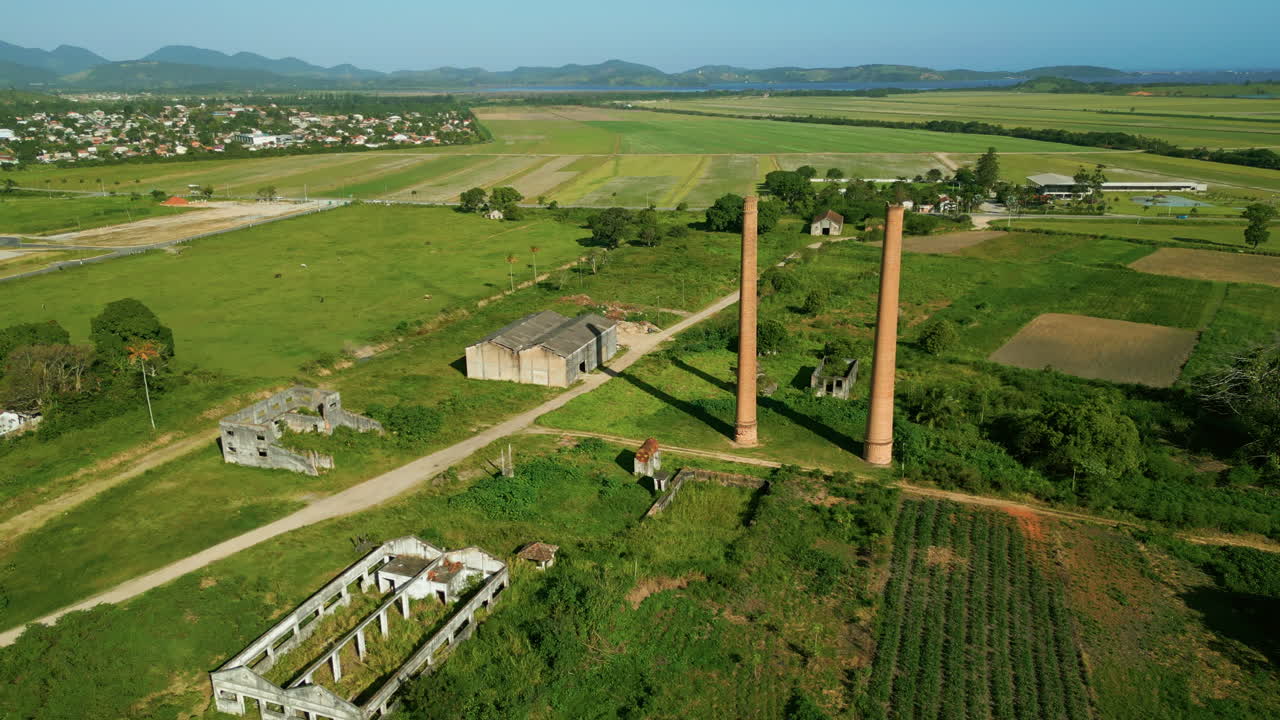 Pengambilan gambar sinematik dengan drone: pengambilan yang menjauh dari pabrik gula dan alkohol yang terbengkalai di Sampaio Correa. Cerobong asap dan reruntuhan bersejarah dikelilingi oleh lanskap pedesaan hijau di Saquarema, Rio de Janeiro. 4K