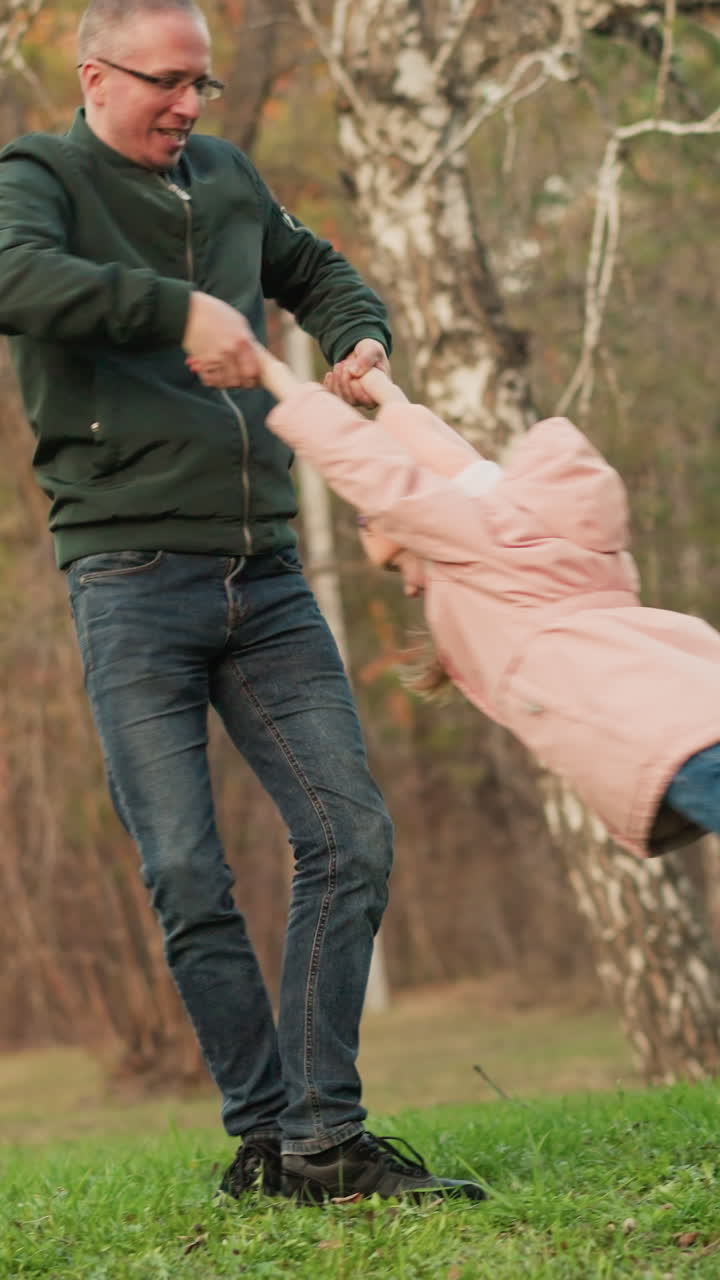 un momento alegre capturado cuando un hombre con una chaqueta verde y pantalones vaqueros azules gira una niña en una chaqueta rosa en un campo verde exuberante en un parque. la escena encarna la felicidad, la unión familiar