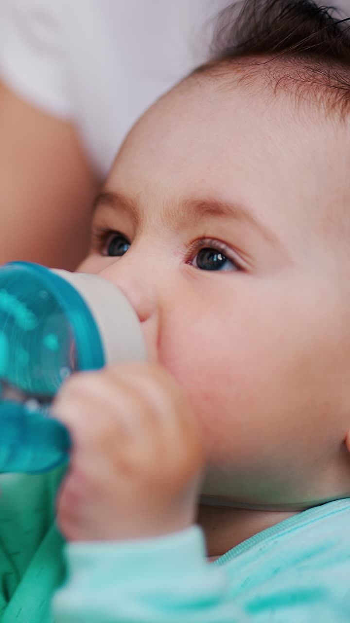 Un adorabile bambino siede tra le braccia della mamma tenendo un biberon. Un dolce bambino caucasico beve acqua. Primo piano. Video verticale