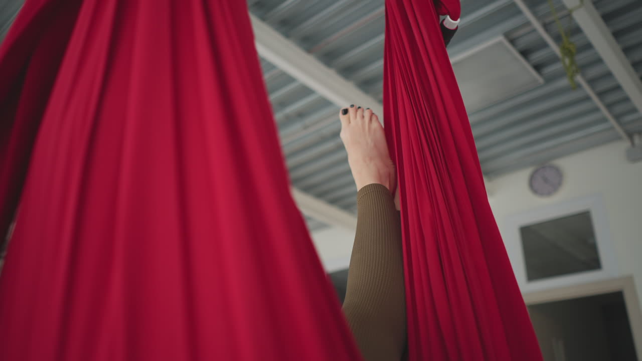 High angle view of performer leg hanging in red silk hammock swinging gently in bright yoga studio with sheer curtains and urban window view conveying calm wellness balance practice flow