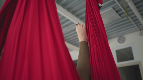 high angle view of performer leg hanging in red silk hammock swinging gently in bright yoga studio with sheer curtains and urban window view conveying calm wellness balance practice flow