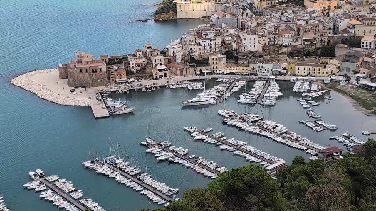 Slow pan up from a hillside viewpoint overlooking Castellammare del Golfo’s marina, castle, and historic old town on the Sicilian coast. Clear daylight view of boats and waterfront architecture