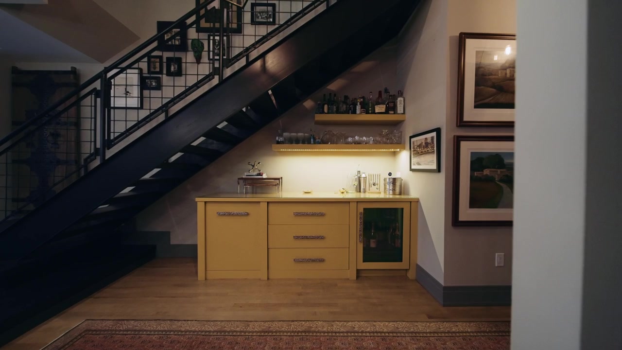 Built-in home bar with yellow cabinetry and open shelving tucked under a modern staircase, featuring soft lighting and a cozy, stylish ambiance