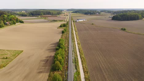 high altitude tracking aerial drone view of high speed passenger train approaching on long straight railroad with fields and forests on both sides. passing under road bridge.