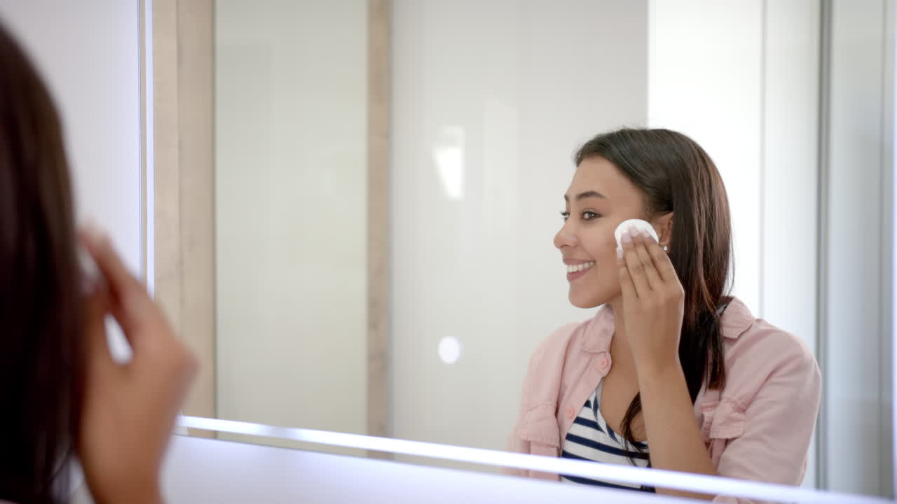 Applying skincare, woman using cotton pad and smiling in bathroom mirror
