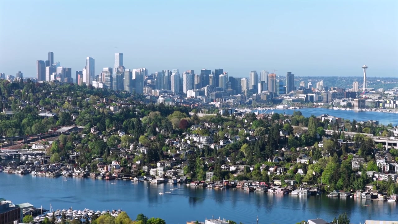 Seattlen siluetti korkeine tornitaloineen Union-järven takana, rehevä asuinalueen rinne ja venesatama, jossa purjeveneitä ankkuroituna tyynessä sinisessä vedessä aurinkoisen taivaan alla. Washington, USA. Laaja ilmapanoraama