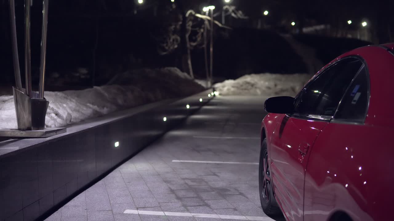 젊은 남자가 밤에 주차장에서 빨간 차에서 내리고 문을 닫습니다. 뒷면.