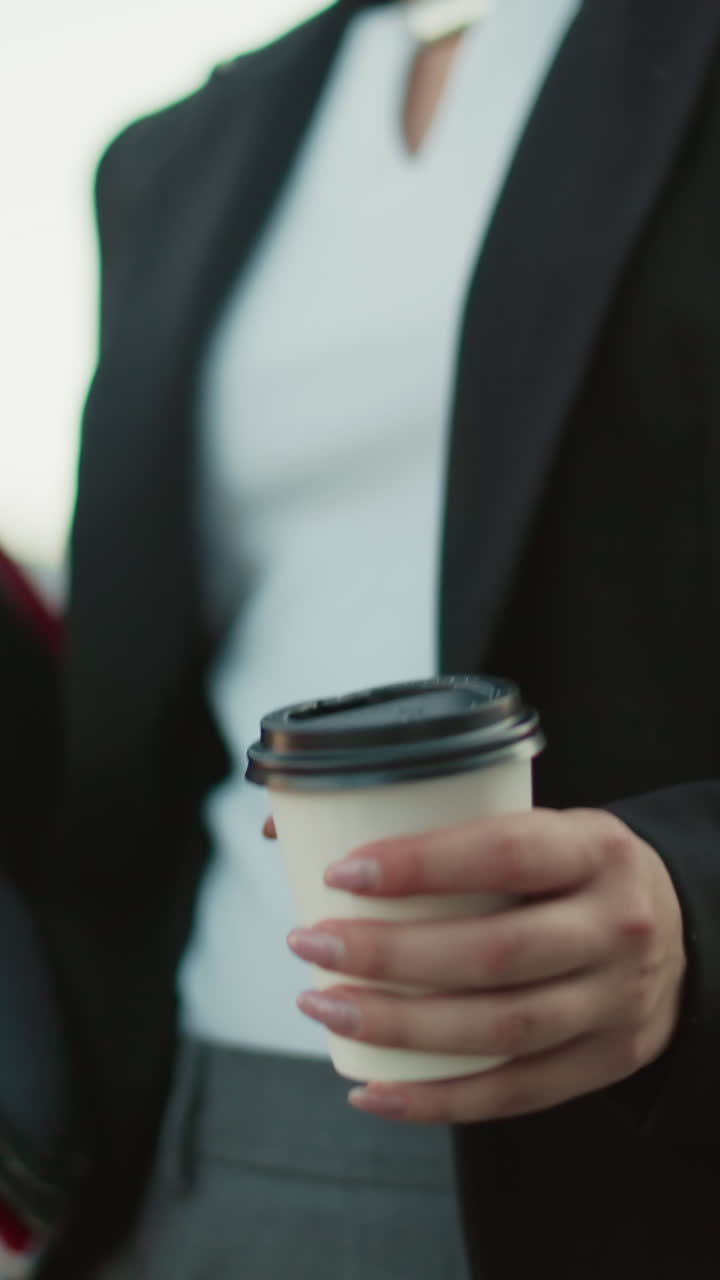 Partial view of office assistant in black suit holding file folder and coffee cup while walking along path with blurred background of parked cars and modern urban buildings