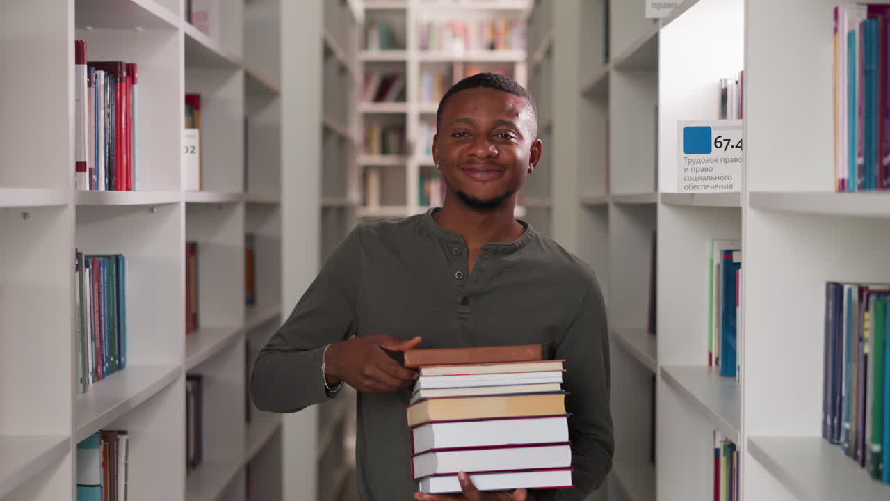 un hombre feliz sostiene una pila de libros en una biblioteca pública. un bibliotecario afroamericano asente con la cabeza llevando una pila de literatura entre las estanterías. un estudiante muestra folio