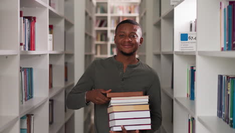 happy man holds book stack in public library. african american librarian nods head carrying literature pile between bookcases. student shows folios