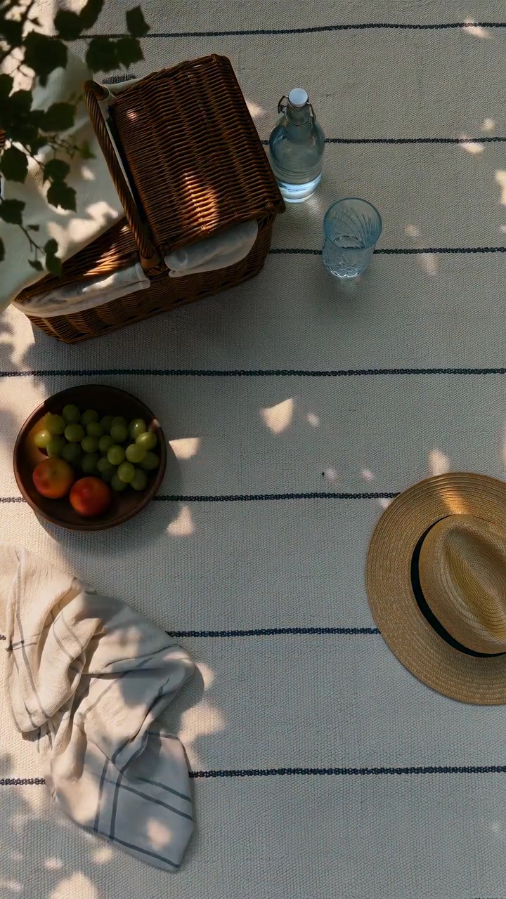 Disposizione da picnic dall'alto che mostra la luce solare e le ombre del fogliame che si spostano gradualmente su una leggera coperta tessuta con sottili strisce scure, creando motivi mutevoli attorno a cesto di vimini, bottiglia, bicchiere