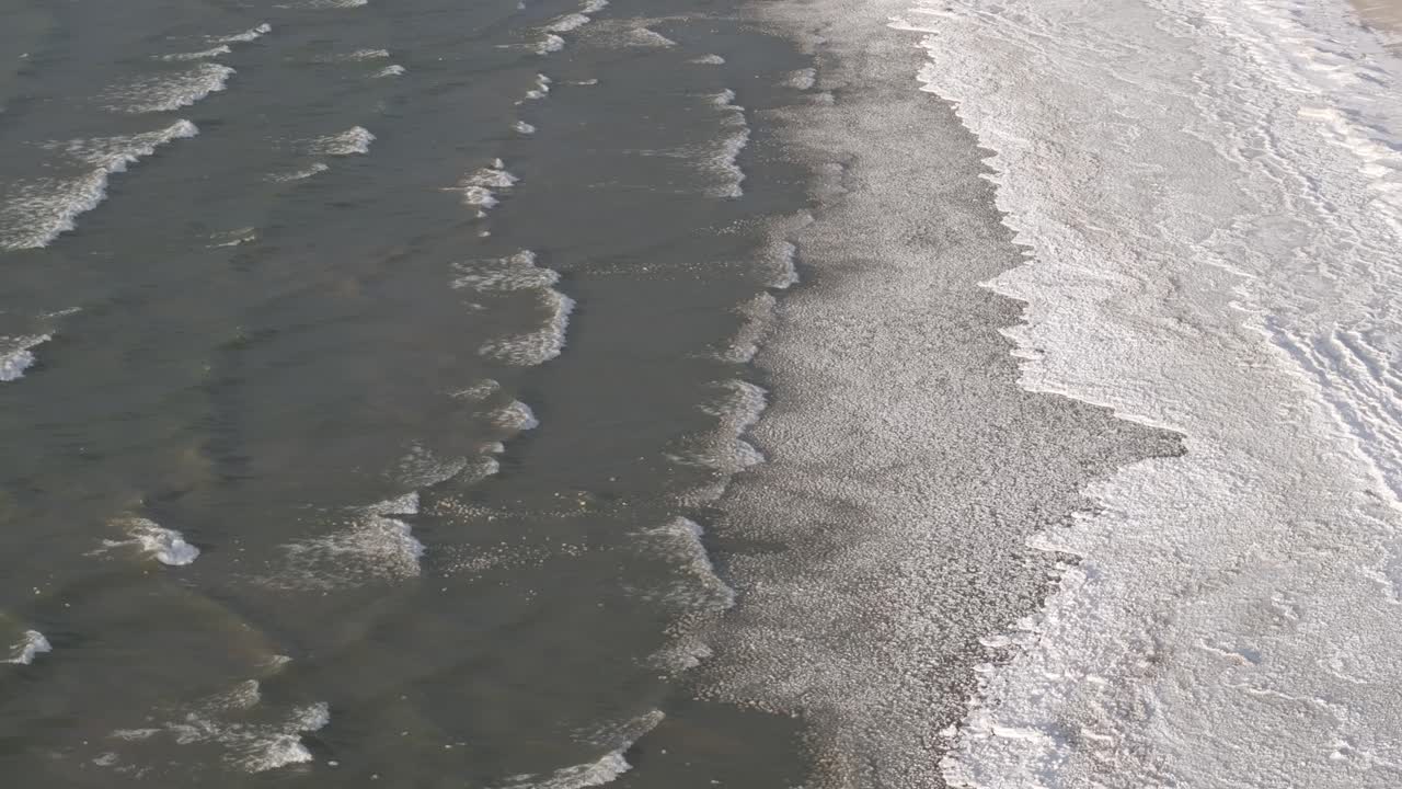 Gentle waves lapping onto a sandy shore, sunlight reflecting on water, aerial view