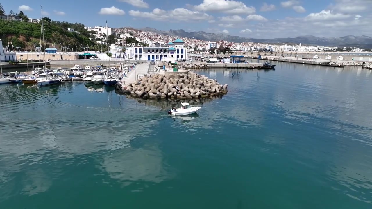 Ampia vista aerea di un porto turistico mediterraneo nel Marocco settentrionale, che mostra yacht ormeggiati, strutture sul lungomare, un bacino portuale protetto e una densa città costiera con montagne sullo sfondo
