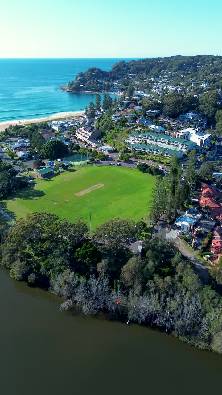 Drone aerial portrait of Avoca Beach local sports fields Heazlett Park with main town shops and cars driving along street with ocean coastline and bushland headland Central Coast tourism Australia