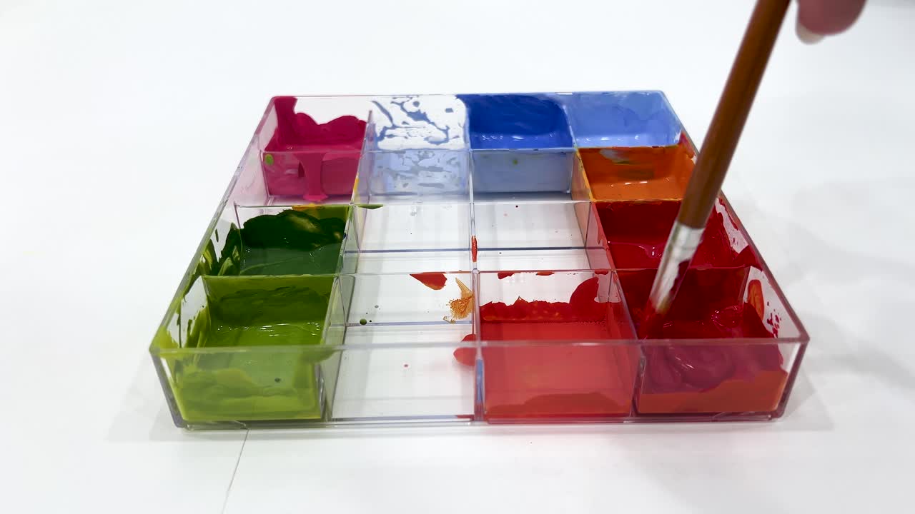 A hand uses a paintbrush to mix red and orange poster paints in a clear plastic tray under bright, even lighting on a white surface