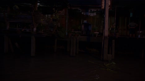 Camera-pans-from-right-to-left,-passing-by-some-houses-on-stilts-in-a-local-community-while-on-a-night-tour-at-Amphawa-Floating-Market-in-Samut-Songkhram,-Thailand