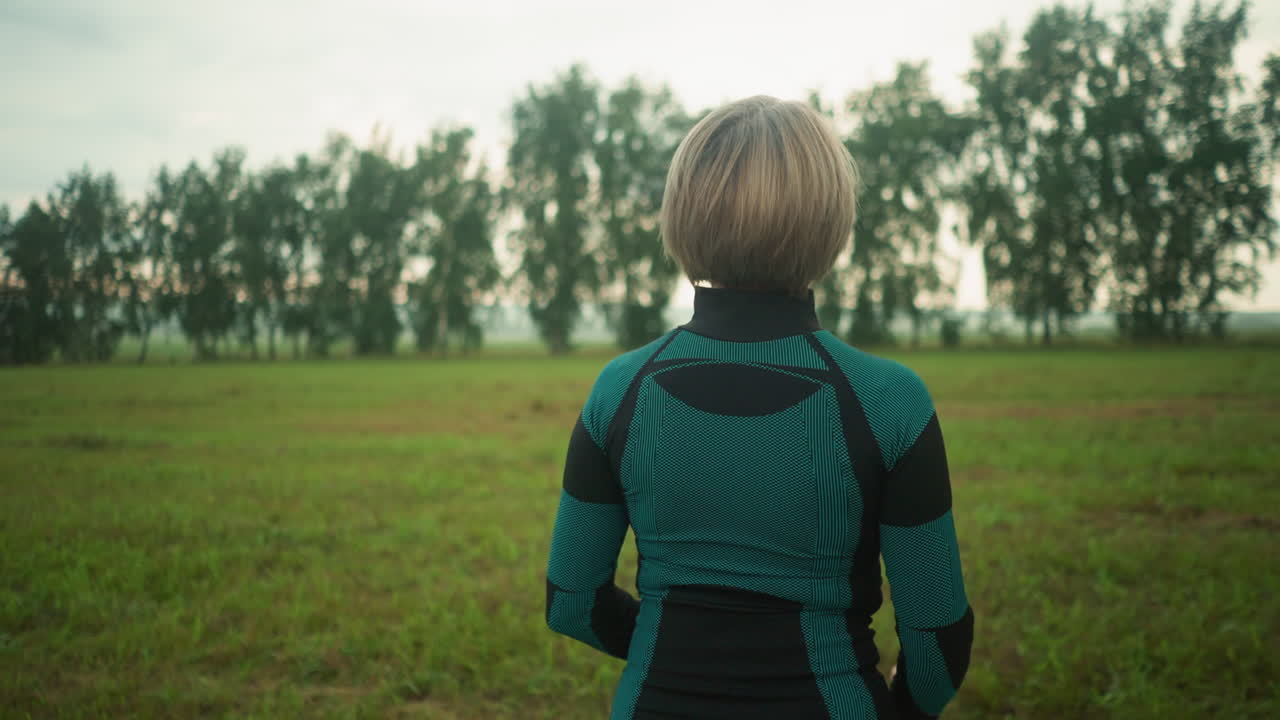 넓은 잔디에서 요가를 연습하는 여성의 뒷면, 멀리 배경에 나무와 함께 조용한 명상에서 손을 천천히 위아래로 움직입니다.