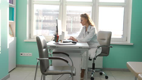 ärztin, die am computer arbeitet, während sie in ihrem sprechzimmer am schreibtisch sitzt