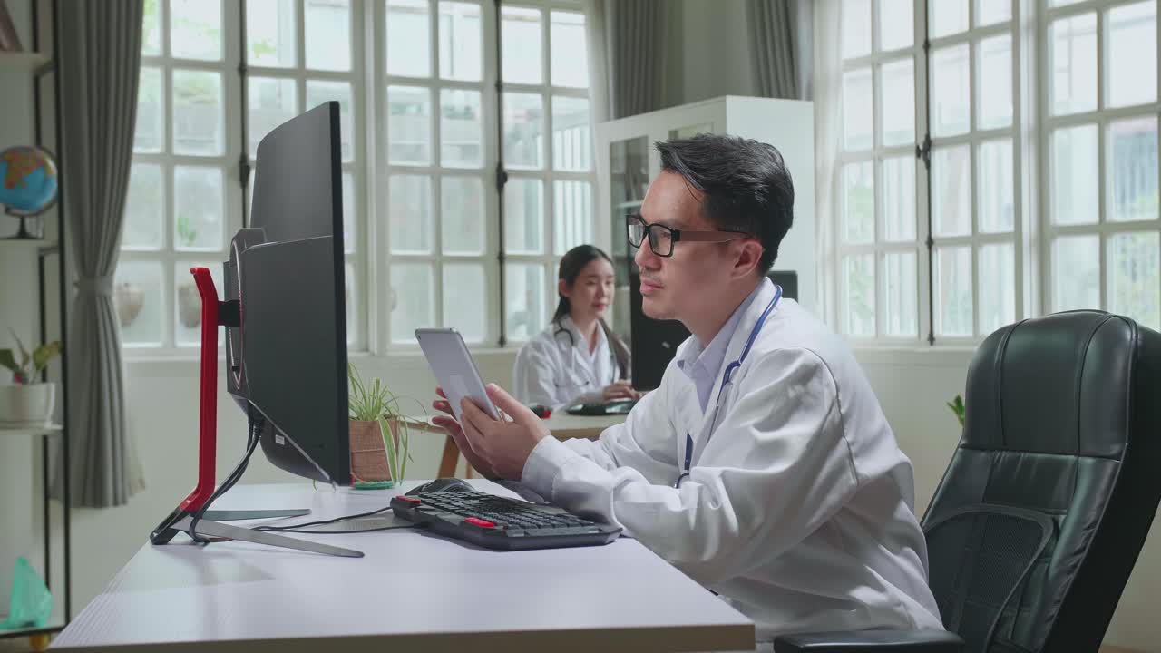 Asian Man Doctor Is Using Tablet While Work With Desktop Computer In Workplace. Medical Concept