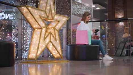 joven sentada en un banco en un centro comercial, deslizando a través de su teléfono con bolsas de compras de colores a su lado y decoraciones festivas, alguien es visible en el fondo en el ascensor