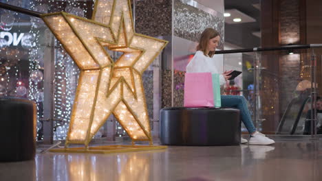 young girl seated on a bench in a shopping mall, sliding through her phone with colorful shopping bags beside her and festive decorations, someone is visible in the background on the elevator