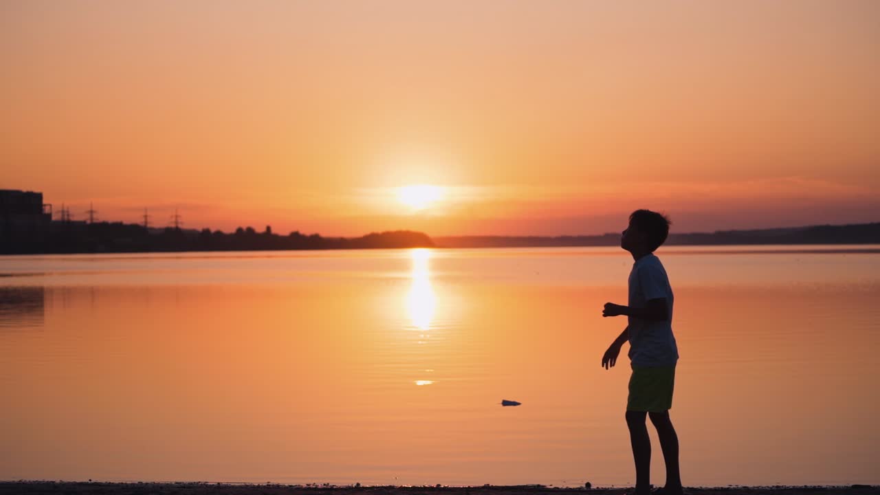 Boy near the river at sunset. Side view of a kid who is throwing paper plane up on the beautiful orange background of calm lake. Slow motion.