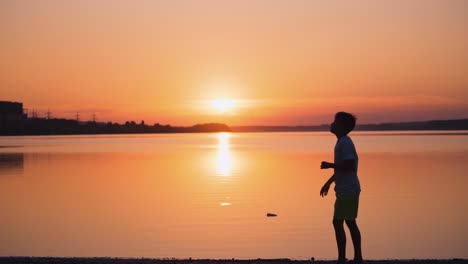 boy near the river at sunset. side view of a kid who is throwing paper plane up on the beautiful orange background of calm lake. slow motion.
