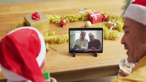 Padre-E-Hijo-Caucásicos-Con-Gorros-De-Papá-Noel-Usando-Una-Tableta-Para-Una-Videollamada-Navideña-Con-La-Familia-En-La-Pantalla