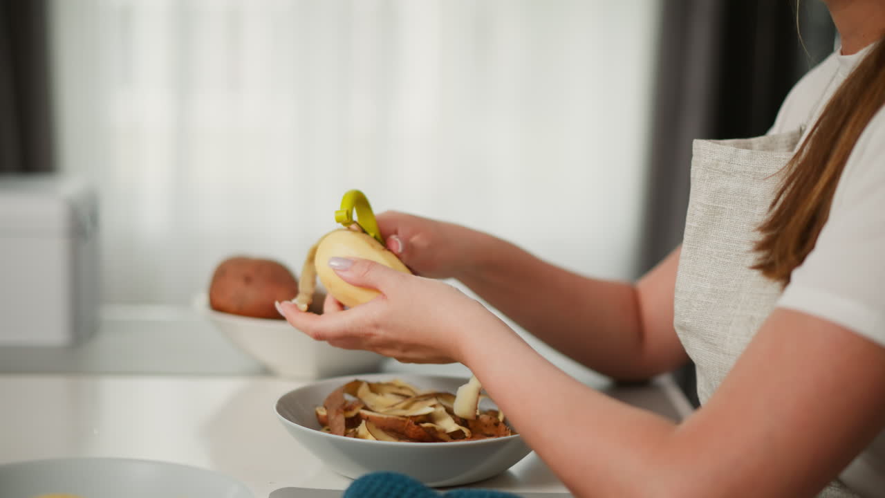 Close up side view of woman peeling potato with green peeler over bowl filled with peels, hair tied back, soft daylight filtering through transparent curtain in calm home kitchen environment