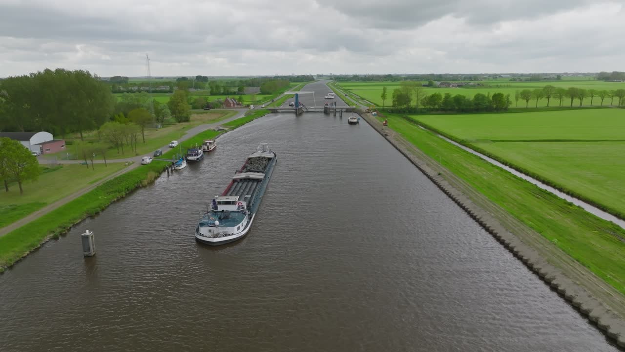 Long overview of a straight Dutch canal with barges and small craft lined up near a rural bascule bridge amid green fields