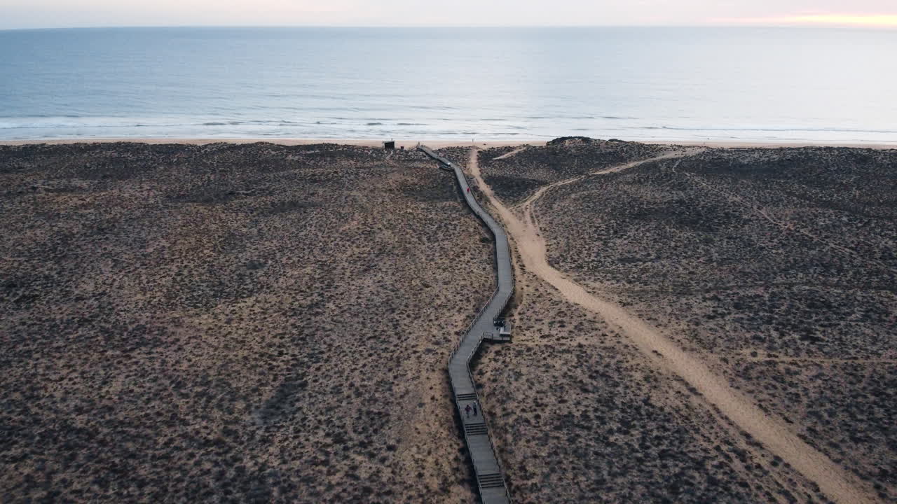 An aerial perspective captures the cinematic beauty of Portugal's coastal landscape, featuring a walking pathway and a solitary road winding through a desolate terrain to water surface