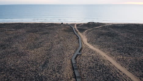 Una-Perspectiva-Aérea-Captura-La-Belleza-Cinematográfica-Del-Paisaje-Costero-De-Portugal,-Presentando-Un-Sendero-Para-Caminar-Y-Un-Camino-Solitario-Que-Serpentea-A-Través-De-Un-Terreno-Desolado-Hasta-La-Superficie-Del-Agua.