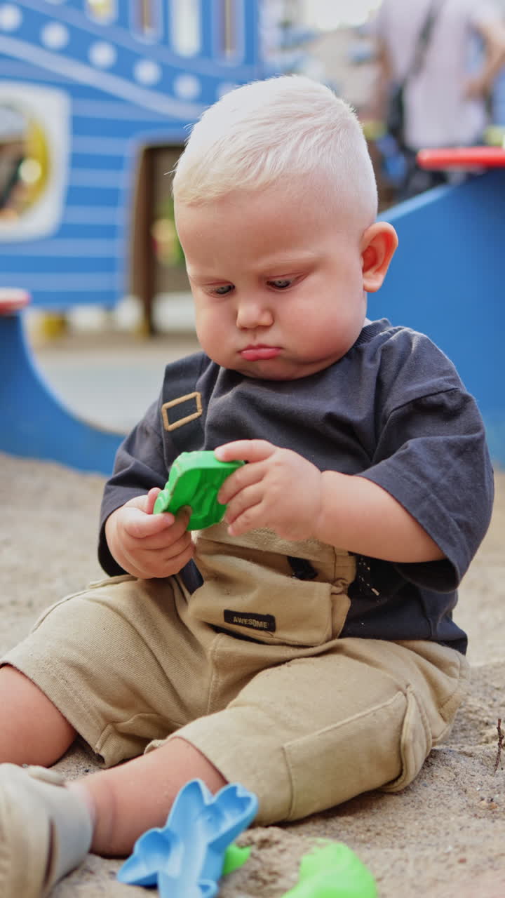 A cute blond boy, 1 year old, is playing on the street. A walk with a baby. Child development. A boy in a t-shirt on a sunny day. Vertical video. Panama on the head