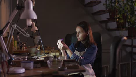 profile of focused caucasian female jeweller sitting at desk, making jewelry in workshop