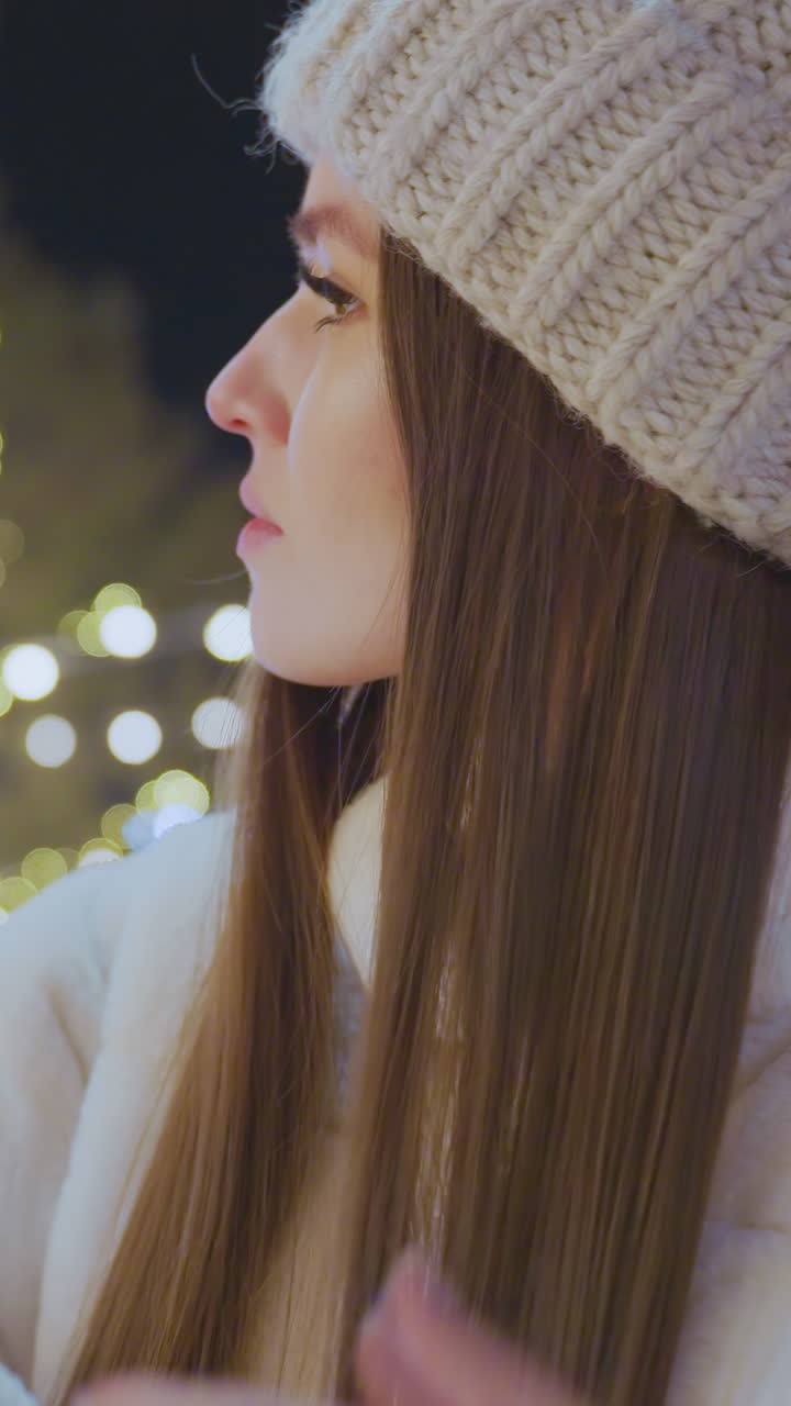 Young woman gently combing her hair outdoors at night, surrounded by glowing decorative lights, city street illuminated with festive bokeh while cars are parked nearby