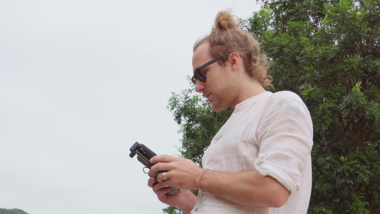 Blond long hair Caucasian man with sunglasses, fly drone with controller, low angle