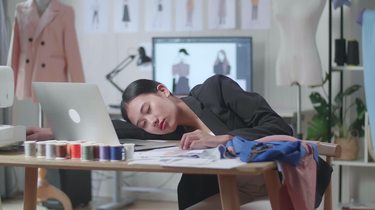 스튜디오에서 옷을 디자인하는 동안 노트북과 함께 비즈니스 슈트를 입은 아시아 여성 디자이너가 잠을 자고 있습니다.